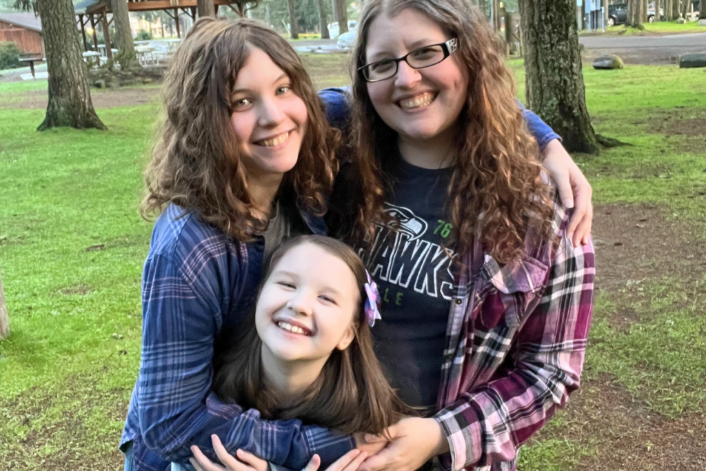 Lacie Ugelstad standing in a local park, surrounded by trees, hugging her two daughters, smiling together.
