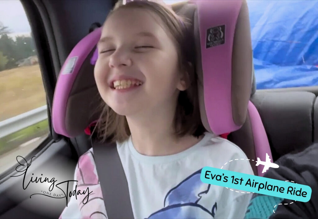 Photo of a young girl smiling in a car seat. Text on the screen says Eva's 1st Airplane Ride along with the Living One Day Today logo